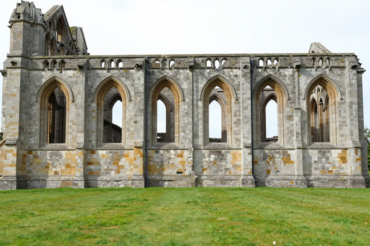A stone ruined abbey with tall empty Gothic arched windows stands beside a green grass field under pale overcast skies
