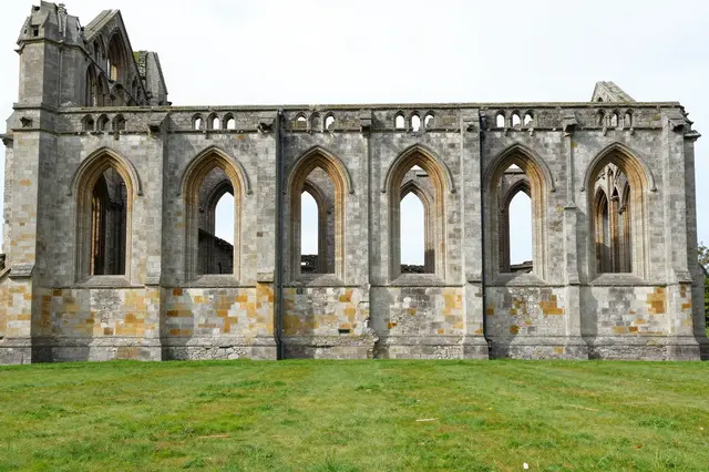 A stone ruined abbey with tall empty Gothic arched windows stands beside a green grass field under pale overcast skies