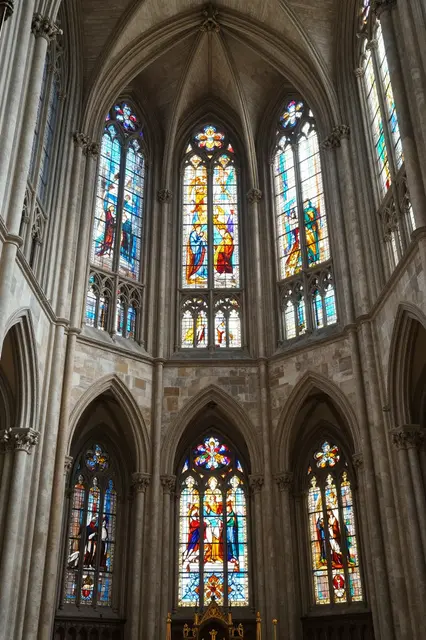 Tall ornate stained glass windows line the stone interior of a gothic cathedral apse