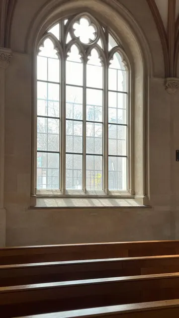 Sunlight streams through a tall arched stone church window onto empty wooden pews