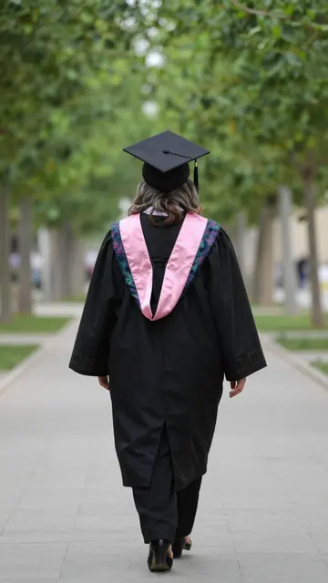 Back view of a woman in graduation regalia walking down a paved tree-lined campus path