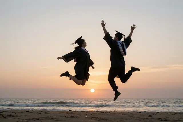 Two graduates in caps and gowns jump in celebration above a sandy beach at golden sunset over the ocean
