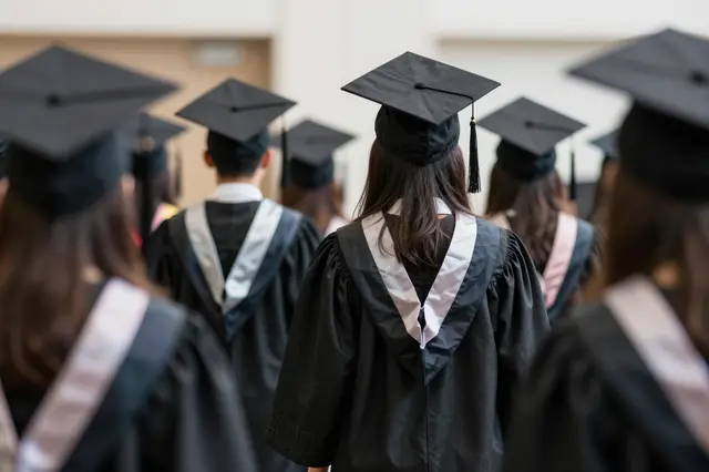 Back view of graduates in black caps and gowns walking toward an indoor graduation ceremony.
