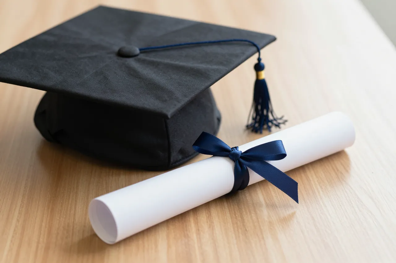 Black graduation cap and rolled diploma tied with navy ribbon rest on a light wooden table