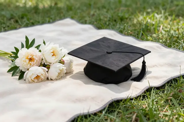 A black graduation cap sits next to a white peony bouquet on a picnic blanket in a sunny grassy park