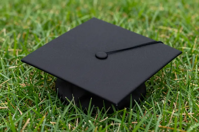 A black graduation mortarboard cap rests on top of lush green lawn grass.