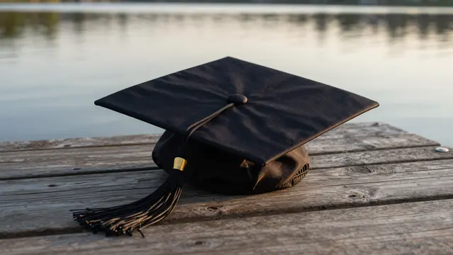 A black graduation cap rests on weathered wooden planks beside a calm lake at golden sunset