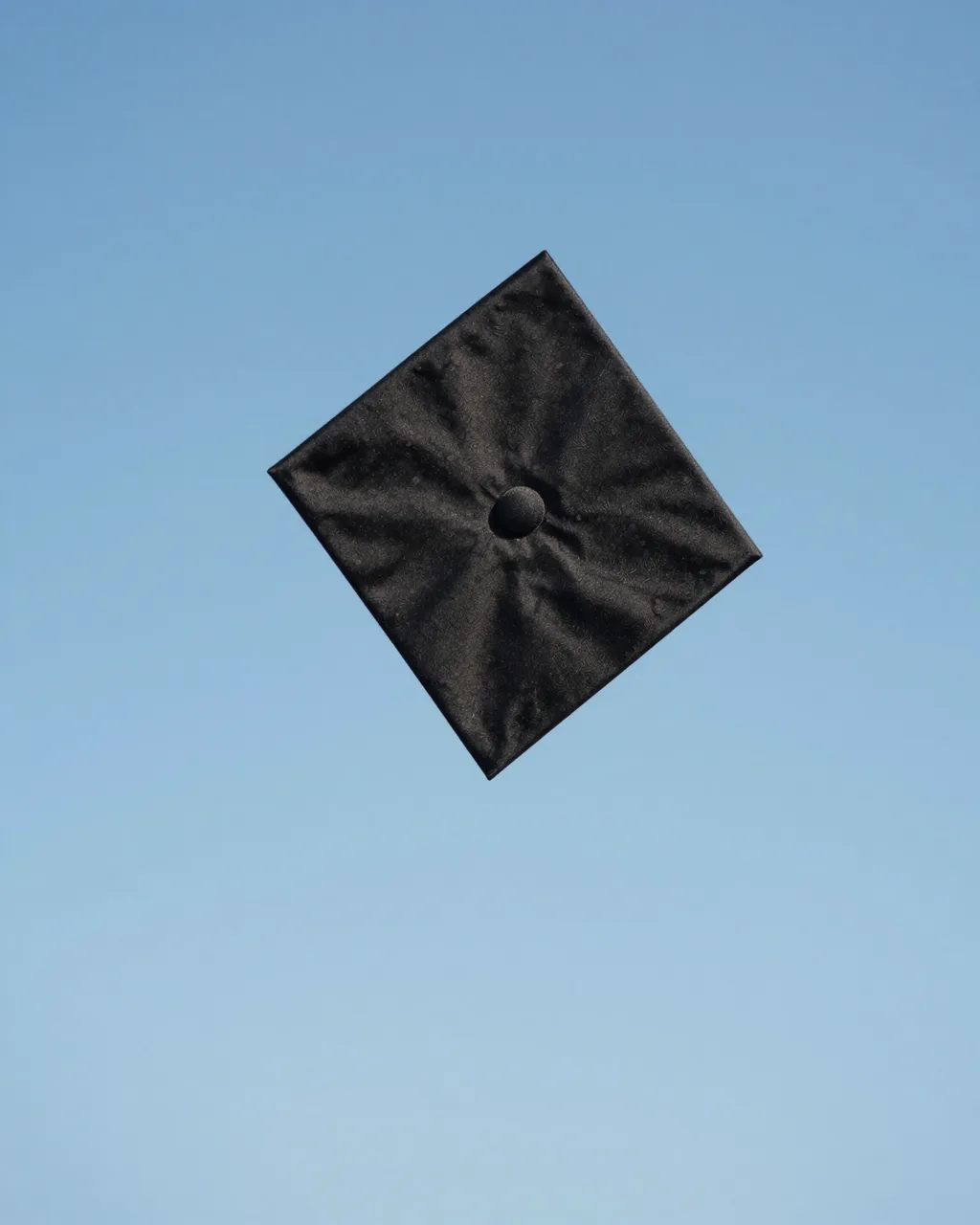 A black graduation cap floats high against a clear light blue sky, viewed from below.