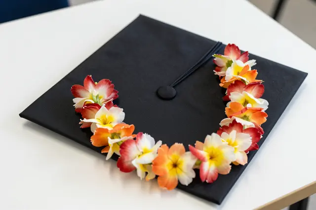 A black graduation cap rests on a white table draped with a colorful flower lei.