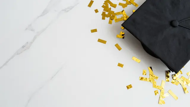 Black graduation cap and scattered gold confetti rest on the right of a white marble surface with empty copy space on the