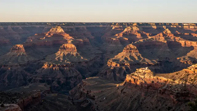 Wide elevated view of layered canyon rock formations glowing in warm sunset light under a pale sky