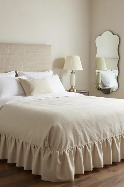 A tidy grandmillennial style bedroom with a ruffled bed skirt, pleated lamp, and scalloped wall mirror