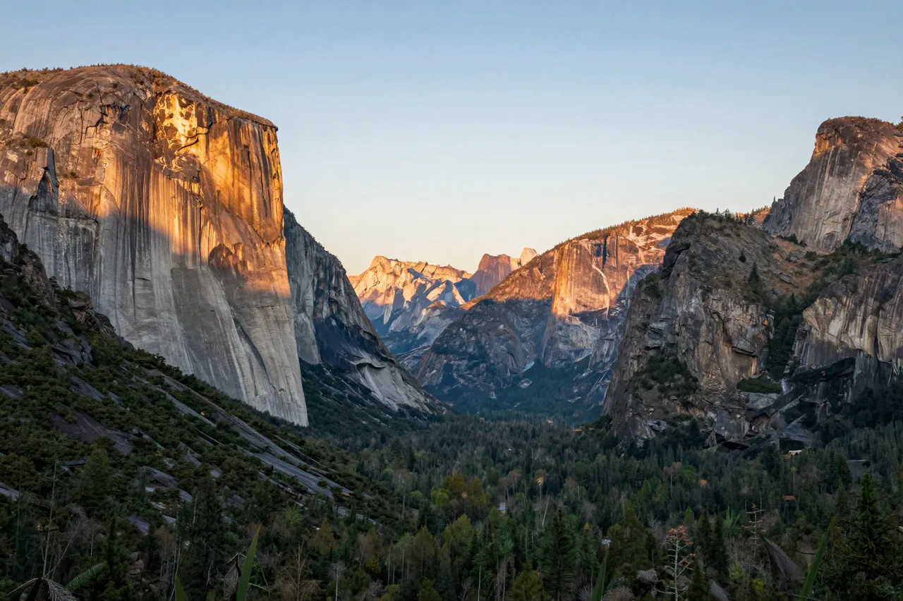 Sheer granite cliff faces glow with warm sunset light above a forested green mountain valley