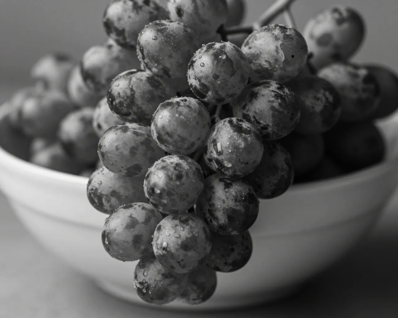 Black and white close-up of a bunch of dewy grapes hanging over the edge of a plain ceramic bowl.