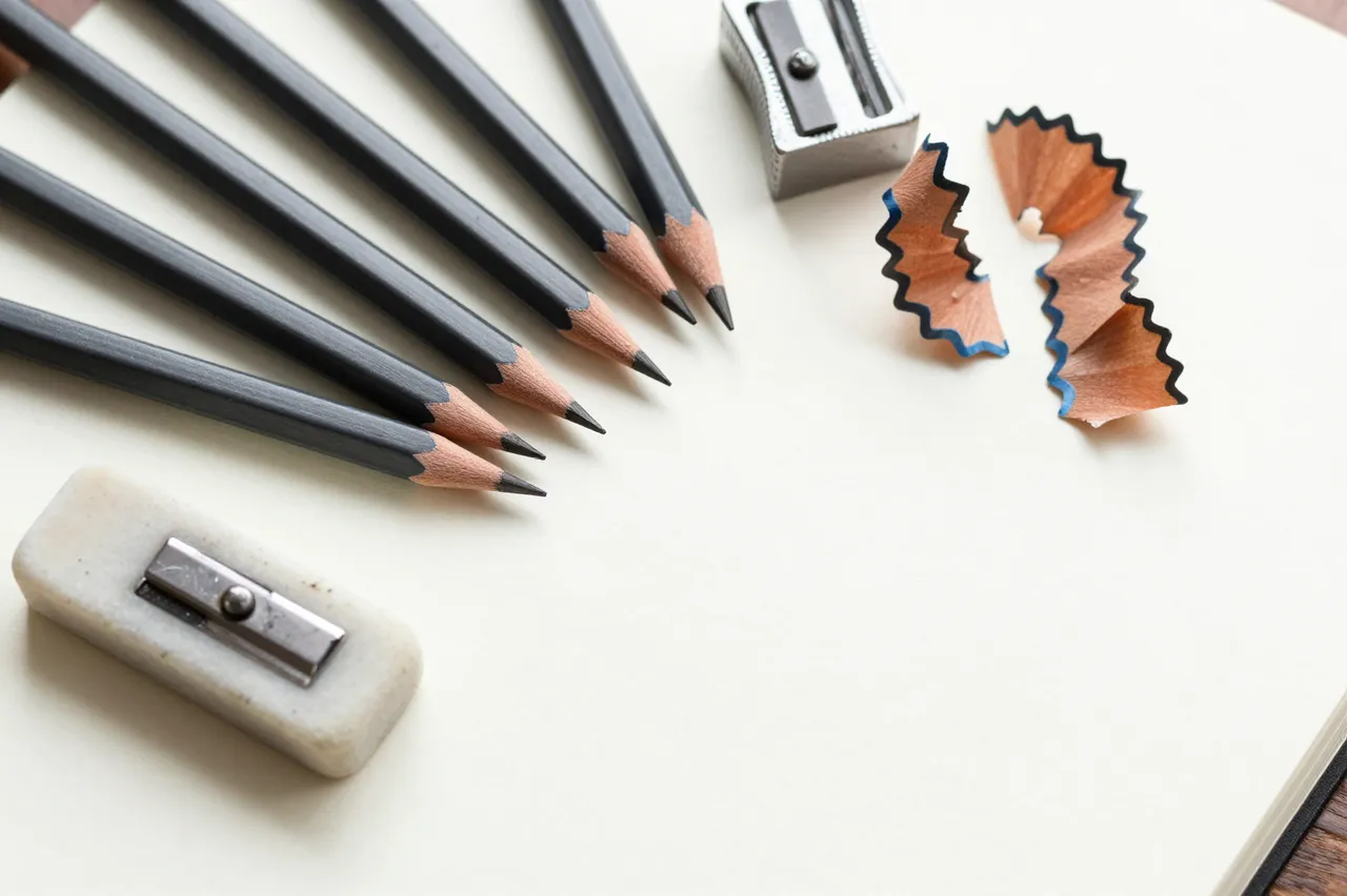 Flat lay arrangement of sharpened graphite pencils, sharpeners, and curled shavings on blank sketchbook paper