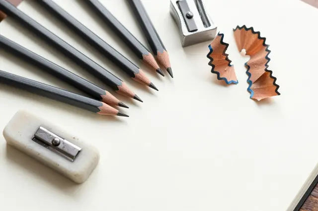 Flat lay arrangement of sharpened graphite pencils, sharpeners, and curled shavings on blank sketchbook paper