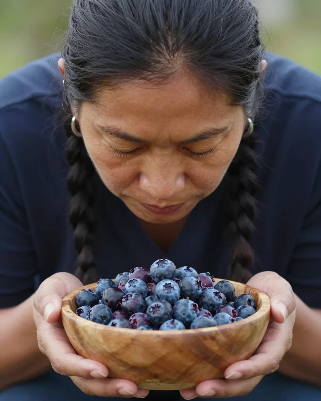 An Indigenous woman with closed eyes holds a wooden bowl of fresh blueberries with both hands, bowing her head in gratitude.