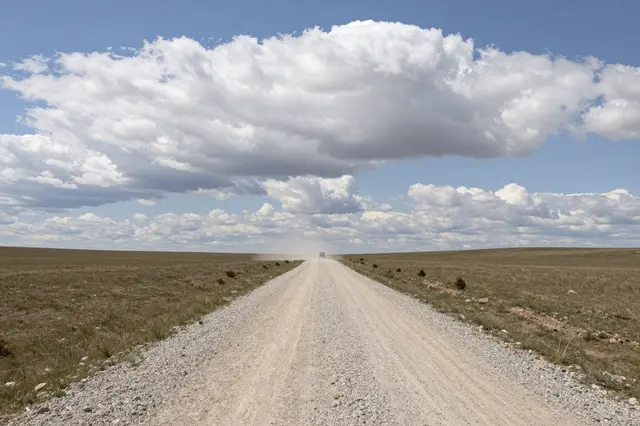 A long gravel steppe road stretches to the horizon where a distant vehicle kicks up dust under large billowing clouds
