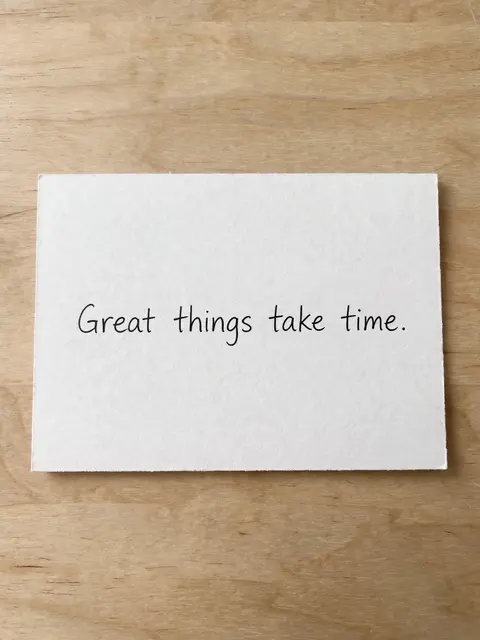 A white index card resting on a light wooden desk displays the handwritten quote "Great things take time."
