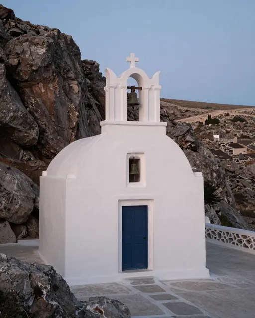 Small white stone chapel with a bell tower set on rocky Greek island terrain at pale dusk
