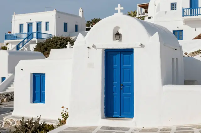Whitewashed Cycladic chapel with bright blue door sits among other white Mediterranean village buildings under clear sky