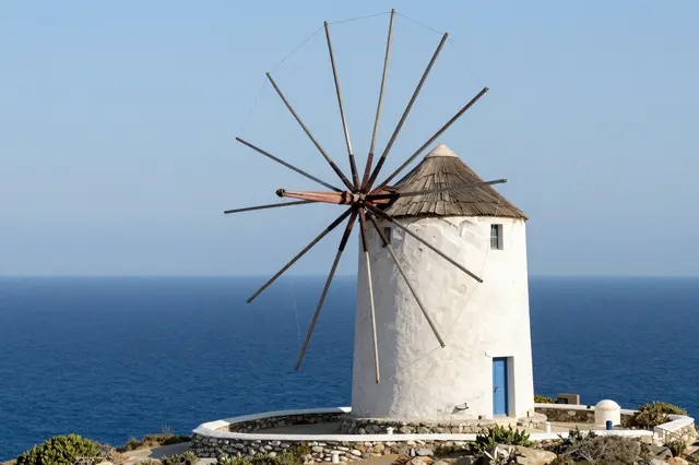 A white stone Greek windmill stands on a coastal hilltop above deep blue sea under clear sky