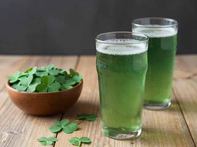 Two glasses of foamy green beer sit next to a wooden bowl of shamrocks on a rustic wooden table.