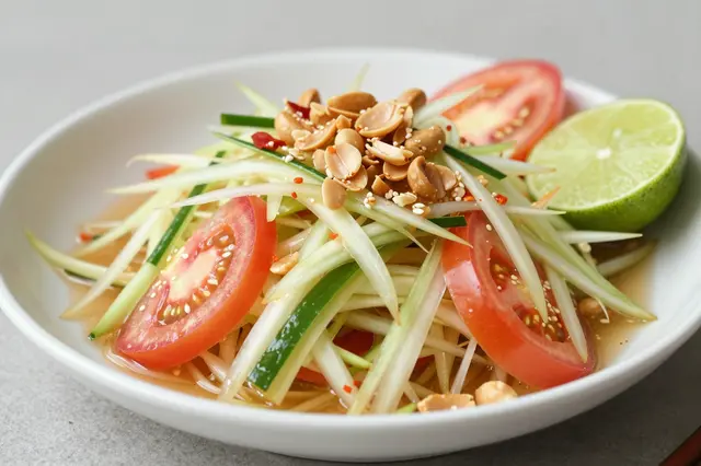Fresh green papaya salad with peanuts, tomato slices and lime served in a white ceramic bowl