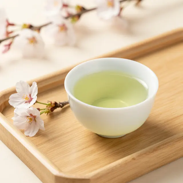 A white bowl of green tea sits on a wooden tray next to a cherry blossom sprig under soft spring light