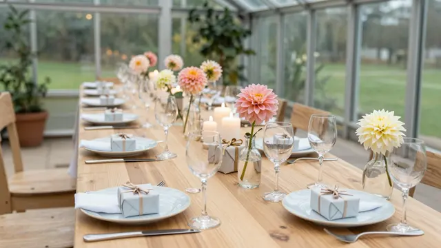 Long wooden dining table set for a celebration inside a glass greenhouse with dahlias, candles and small gifts at each place
