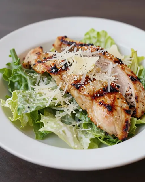 A bowl of grilled chicken caesar salad topped with shaved parmesan on a dark tabletop