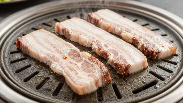 Three slices of pork belly sizzle on a tabletop Korean barbecue grill as faint steam rises.