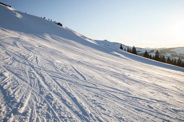 Wide groomed ski slope at golden sunrise, with distant silhouettes along the upper ridgeline