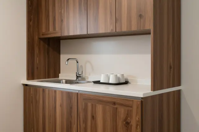 A compact modern kitchenette with walnut cabinets, stainless steel sink, and tray of white cups.