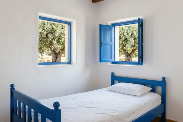 Bright midday guesthouse bedroom with whitewashed walls, blue wooden bed, and olive trees visible through blue-framed windows