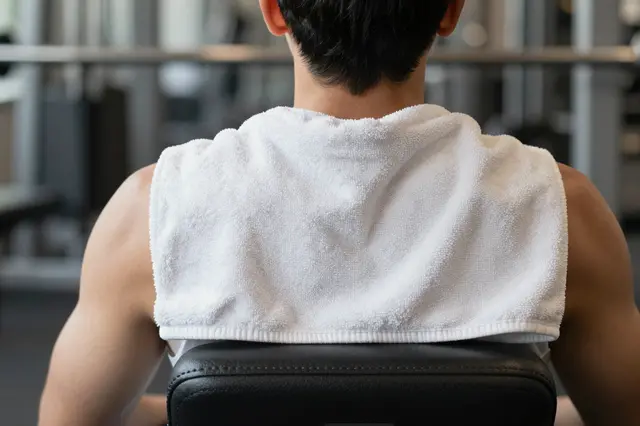 Rear view of an adult resting on a gym bench with a white towel draped over their shoulders in a gym.