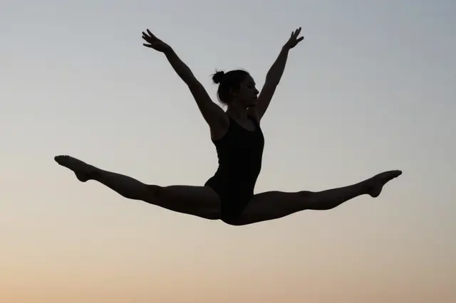 Silhouette of a female gymnast executing a full split leap against a soft pale dawn sky