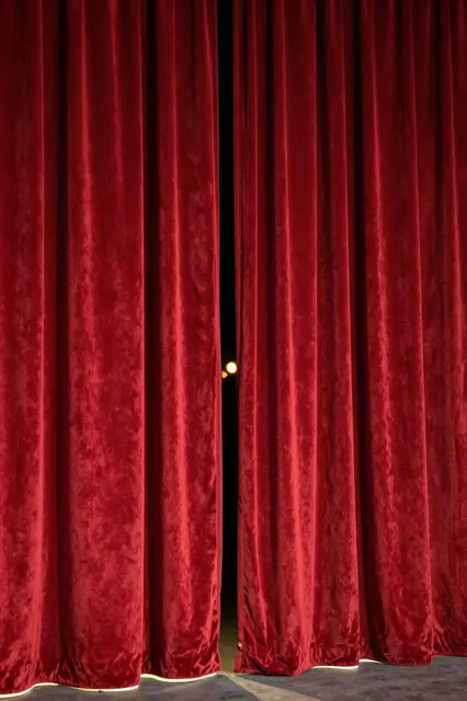 Heavy red velvet stage curtains stand slightly parted with glowing warm footlights at the base