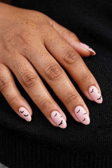 A Pacific Islander woman's hand rests on black knit fabric showing almond pink nails with tiny black bat decals