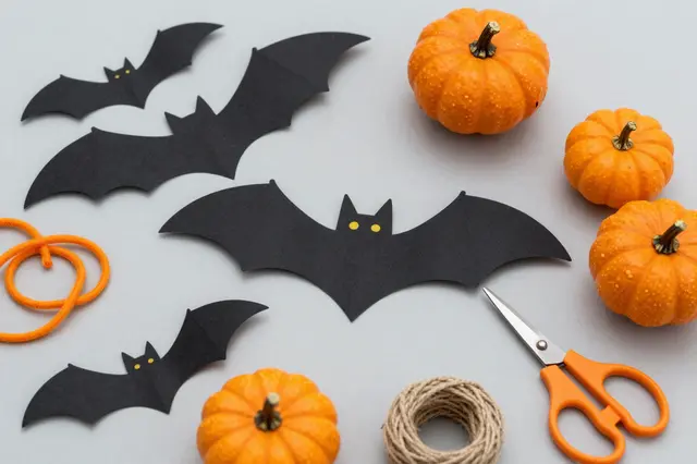 Flat lay of Halloween craft materials including paper bats, mini pumpkins, yarn, twine and scissors on a pale gray surface.