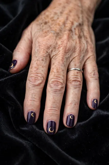 Close up of an older woman's hand with dark purple crescent moon nails resting on black velvet fabric