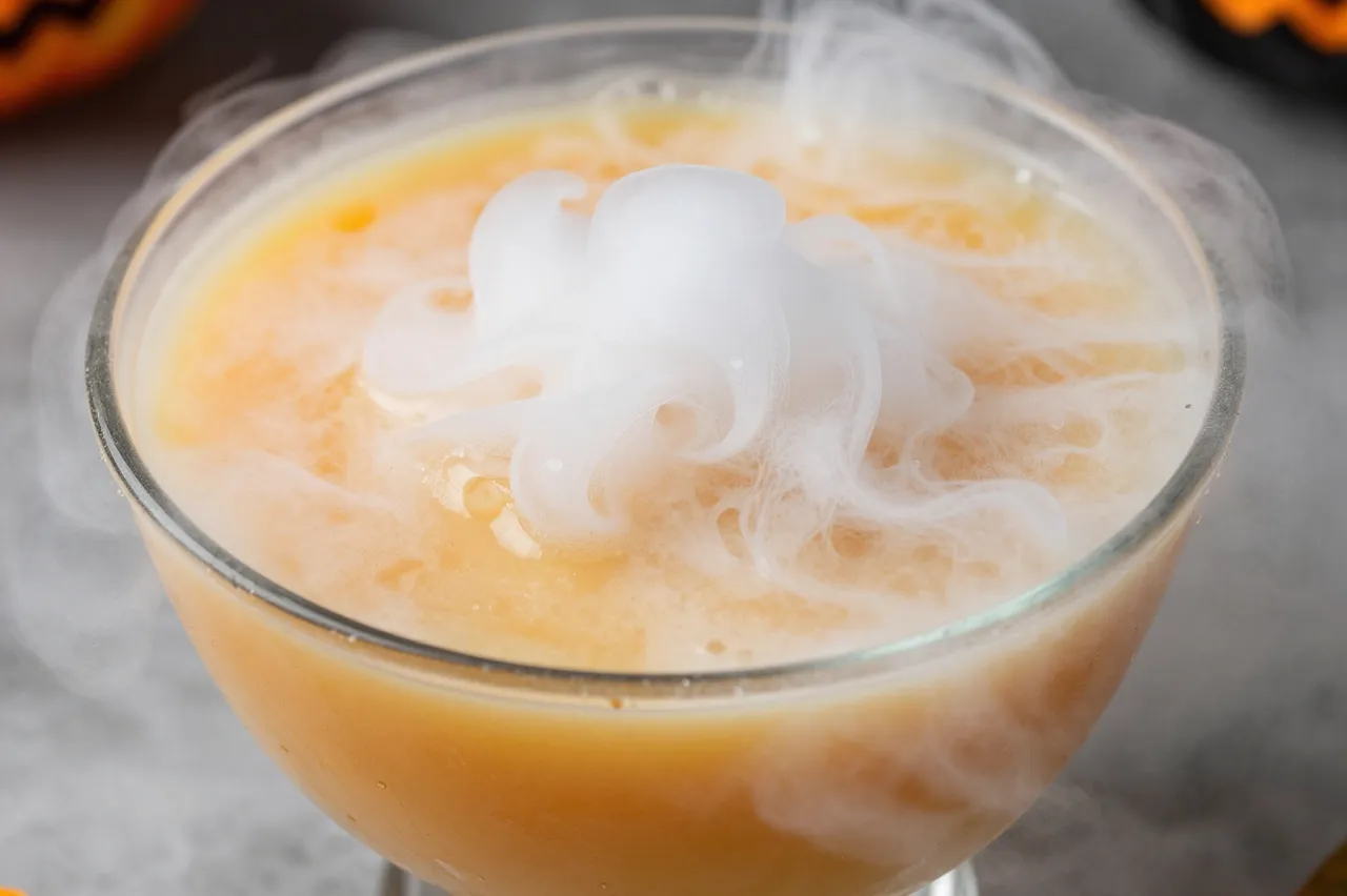 A close-up of a glass bowl of orange Halloween punch wisping with dry ice fog.