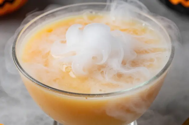 A close-up of a glass bowl of orange Halloween punch wisping with dry ice fog.