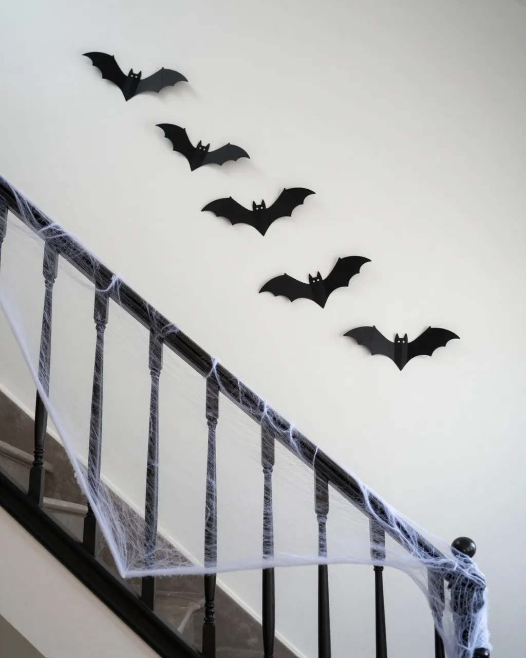 Black paper bat decals line a white stair wall above a banister wrapped in faux cobwebs