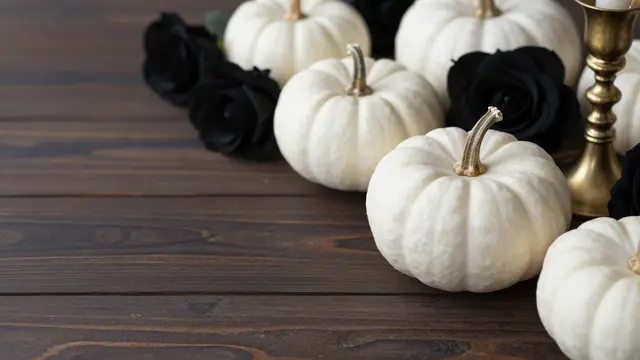 White pumpkins, black roses, and a brass candlestick sit on the right of a dark wooden table with empty left copy space