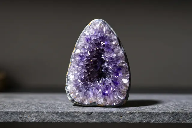 A cut amethyst geode stands upright on a dark textured stone shelf lit by soft side light.