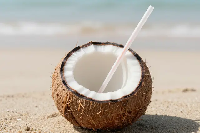 A halved fresh coconut with a drinking straw rests on sunlit beach sand near soft out-of-focus ocean waves.