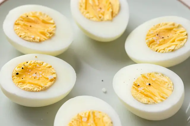 Six halved hard boiled eggs sprinkled with flaky salt and pepper rest on a pale ceramic plate