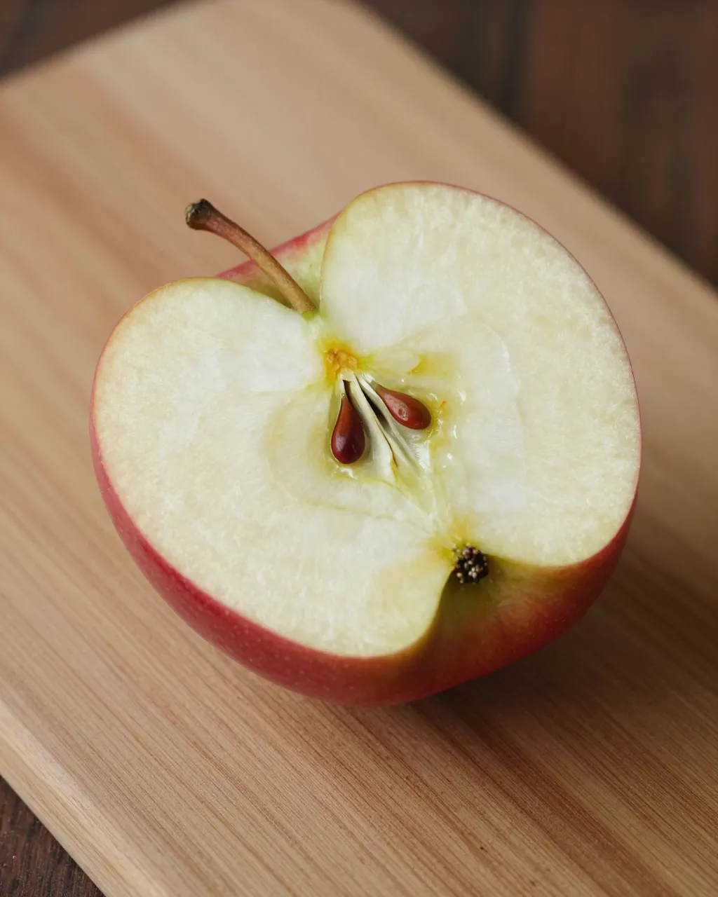 A halved red apple with visible seeds rests on a light wooden cutting board.