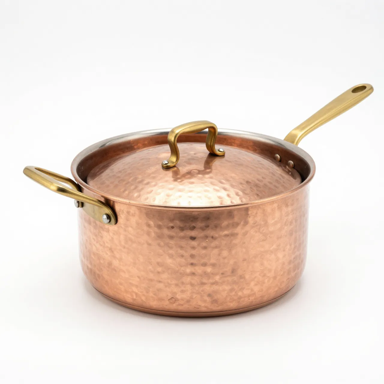 Hammered copper saucepan with brass handles and lid sits isolated on a plain white studio background